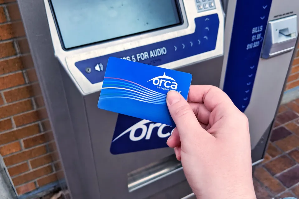 Close-up of person's hand holding blue ORCA transit fare payment card in front of fare payment machine.